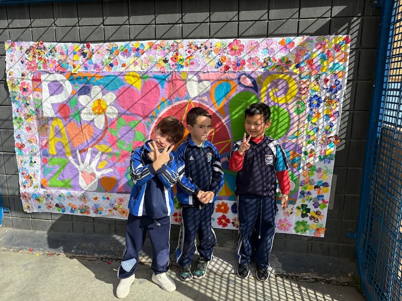 Tres alumnos sonriendo y haciendo el símbolo de la paz frente al mural del colegio en el Día de la Paz.